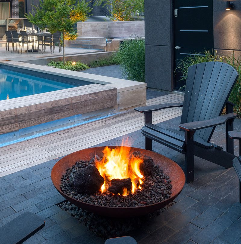 A modern backyard setting with a bowl-shaped fire pit, wooden seating, and a swimming pool in the background, during the evening.